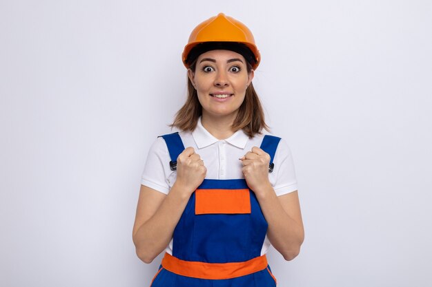 Jeune femme de construction en uniforme de construction et casque de sécurité heureux et excité debout sur un mur blanc