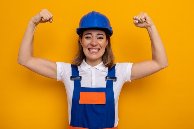 Jeune femme de construction en uniforme de construction et casque de sécurité heureux et excité levant les poings comme un gagnant debout sur un mur orange