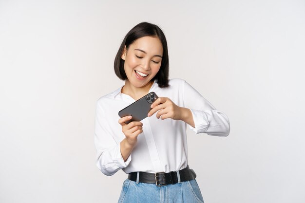 Jeune femme coréenne fille asiatique jouant au jeu vidéo mobile sur smartphone regardant l'écran du téléphone horizontal debout sur fond blanc
