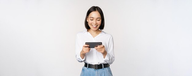 Jeune femme coréenne fille asiatique jouant au jeu vidéo mobile sur smartphone regardant l'écran du téléphone horizontal debout sur fond blanc