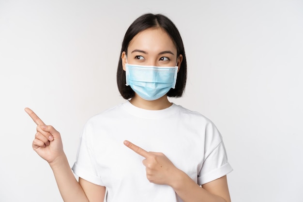 Jeune femme coréenne en masque médical pointant les doigts vers la gauche et regardant le logo montrant une publicité ou une bannière debout sur fond blanc