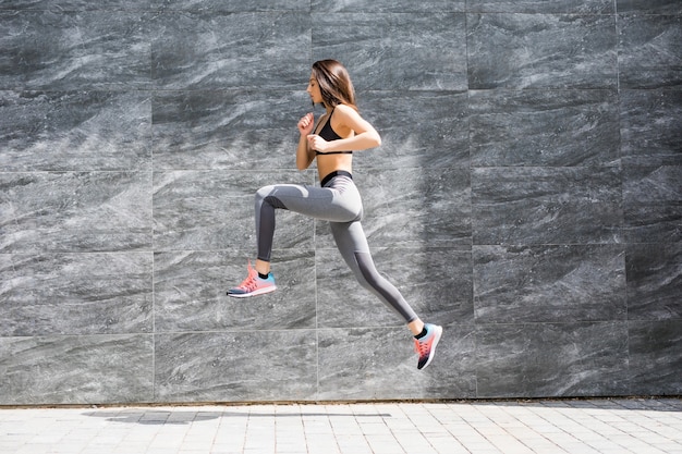 Jeune femme avec corps en forme sautant et en cours d'exécution contre le mur gris.