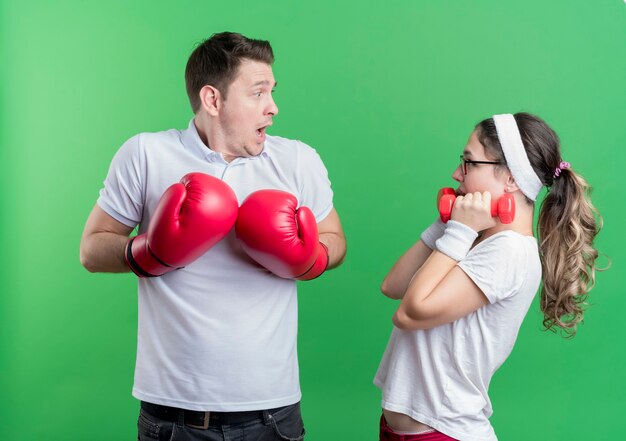 Jeune femme couple sportif avec des haltères en regardant son petit ami avec des gants de boxe surpris et confus debout sur le mur vert