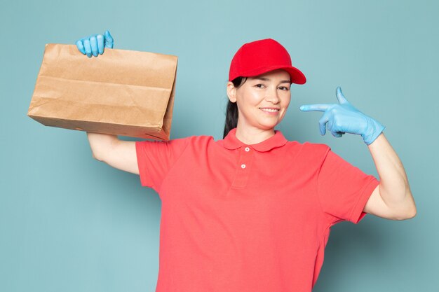 jeune femme courrier en rose t-shirt chapeau rouge gants bleus tenant la boîte sur le mur bleu