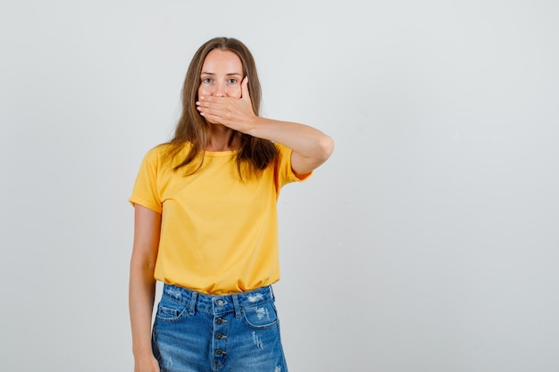 Jeune femme couvrant la bouche avec la main en t-shirt, short et à la recherche de silence. vue de face.