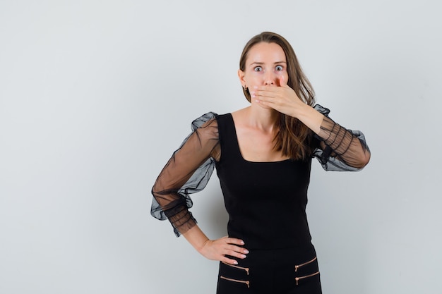 Jeune femme couvrant la bouche d'une main et tenant une autre main sur la taille en chemisier noir et pantalon noir et à la choqué. vue de face.
