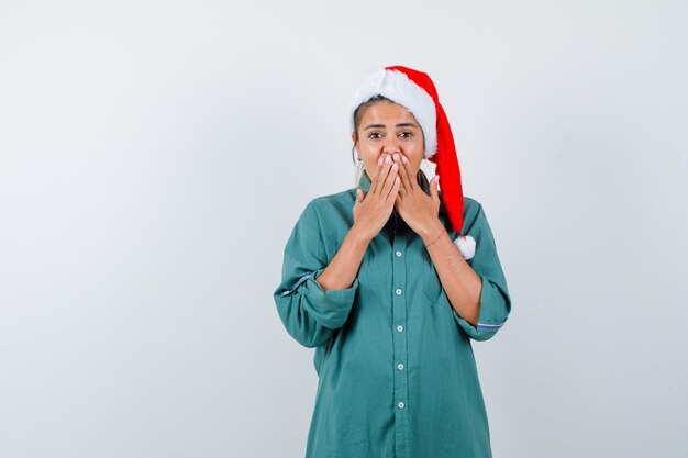 Jeune femme couvrant la bouche avec les mains en chemise, bonnet de Noel et l'air étonné, vue de face.