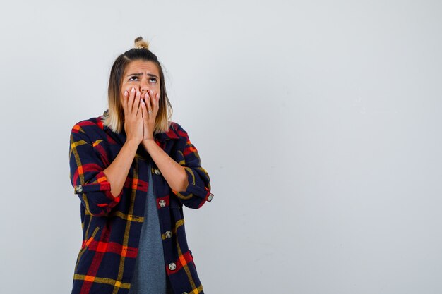 Jeune femme couvrant la bouche avec les mains en chemise à carreaux décontractée et l'air bouleversée, vue de face.