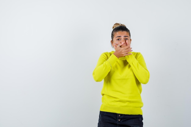 Jeune femme couvrant la bouche avec les mains en pull jaune et pantalon noir et à l'air excité