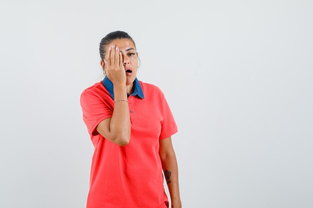 Jeune femme couvrant une partie du visage avec la main en t-shirt rouge et à la vue fatiguée, de face.