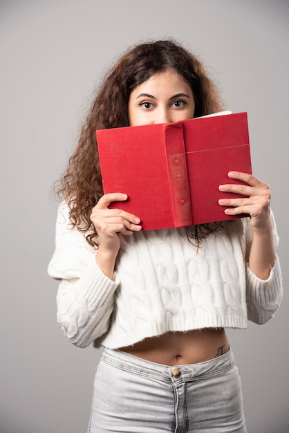 Jeune femme couvrant son visage avec un livre rouge sur un mur gris. Photo de haute qualité