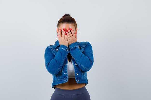 Jeune femme couvrant le visage avec les mains en haut, veste en jean et l'air triste. vue de face.