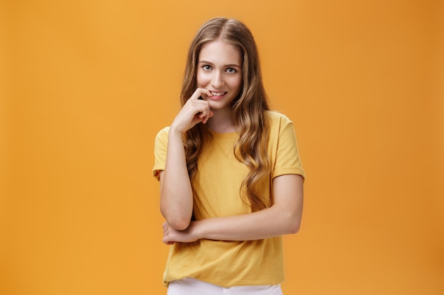 Jeune femme créative et délicate aux cheveux longs naturels ondulés en t-shirt jaune regardant sous le front avec intention et convoitise dans l'expression mordant le doigt, souriant à la caméra sur un mur orange.
