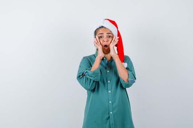 Jeune femme criant en gardant les mains près de la bouche en chemise, bonnet de Noel et l'air troublé, vue de face.