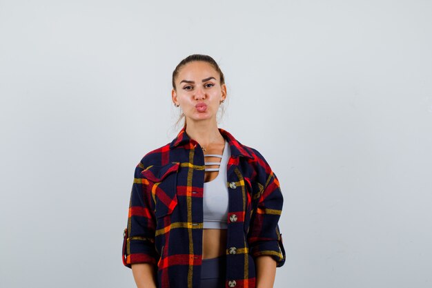 Jeune femme en crop top, chemise à carreaux boudant les lèvres et l'air mignon, vue de face.