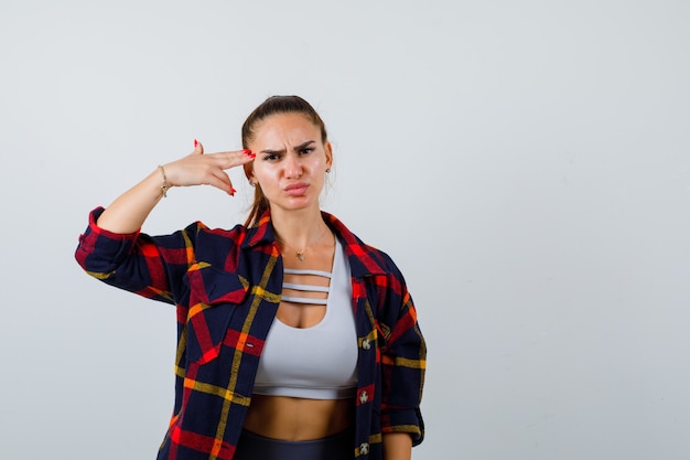 Jeune femme en crop top, chemise à carreaux, pantalon faisant un geste de suicide et semblant sérieux, vue de face.