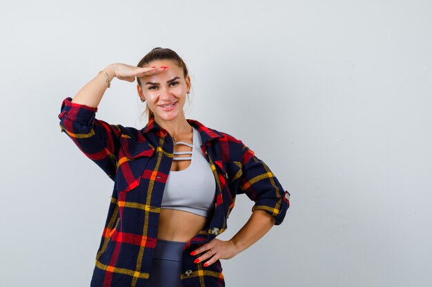 Jeune femme en crop top, chemise à carreaux, pantalon avec la main sur la tête pour voir clairement tout en gardant la main sur la hanche et l'air heureux, vue de face.