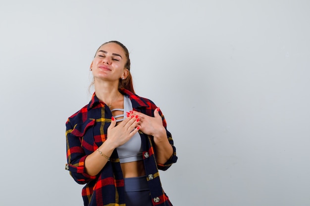 Jeune femme en crop top, chemise à carreaux, pantalon avec les mains sur la poitrine pour prier et avoir l'air plein d'espoir, vue de face.