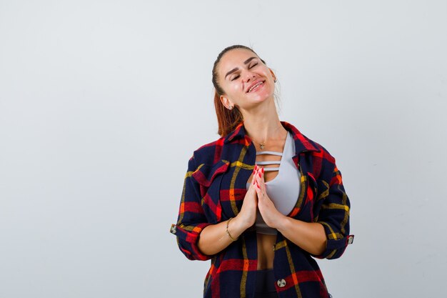 Jeune femme en crop top, chemise à carreaux, pantalon montrant les mains jointes dans un geste implorant et l'air plein d'espoir, vue de face.