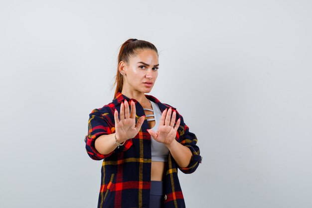 Jeune femme en crop top, chemise à carreaux, pantalon montrant les paumes en geste d'abandon et l'air stressé, vue de face.