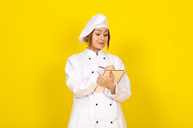 Jeune femme cuisine en costume de cuisinier blanc et bonnet blanc en écrivant des notes