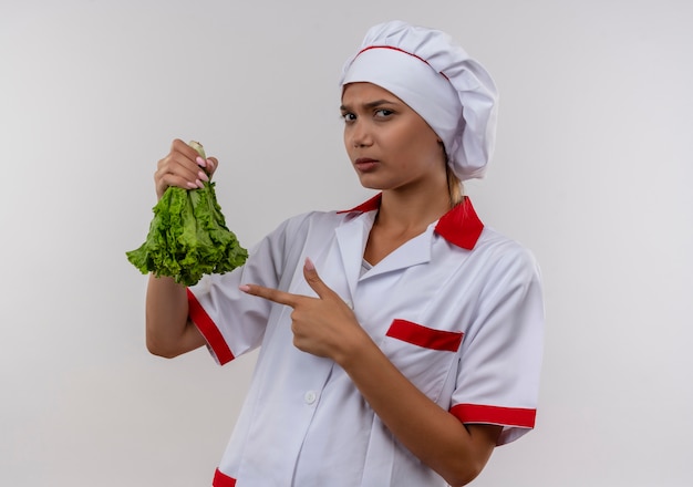 Jeune femme cuisinier portant l'uniforme de chef pointe le doigt à la salade dans sa main sur un mur blanc isolé
