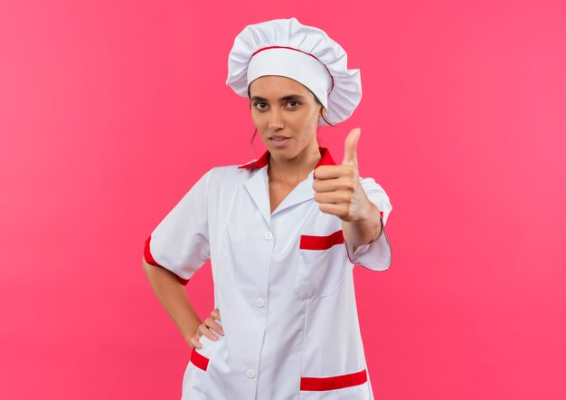 Jeune femme cuisinier portant l'uniforme de chef son pouce vers le haut et mettant la main sur la hanche sur un mur rose isolé avec copie espace