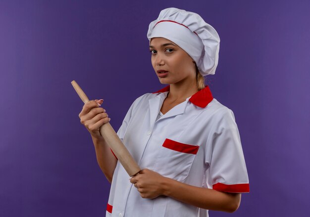 Jeune femme cuisinier portant un uniforme de chef tenant un rouleau à pâtisserie sur un mur isolé