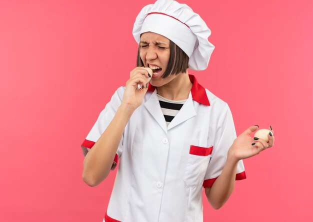 Jeune femme cuisinier en uniforme de chef tenant et essayant de mordre les oeufs isolés sur fond rose avec copie espace