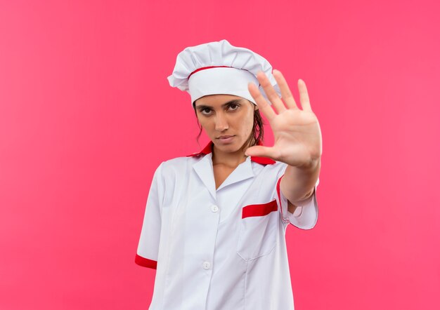 Jeune femme cuisinière portant l'uniforme de chef montrant le geste d'arrêt sur un mur rose isolé avec copie espace