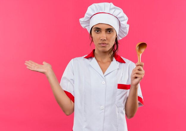 Jeune femme cuisinière portant un uniforme de chef tenant cuillère et points avec la main à l'autre sur un mur rose isolé avec copie espace