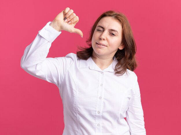 Jeune femme, dans, chemise blanche, regarder front, sourire, et, clignotant, pointant, elle-même, à, pouce, debout, sur, mur rose