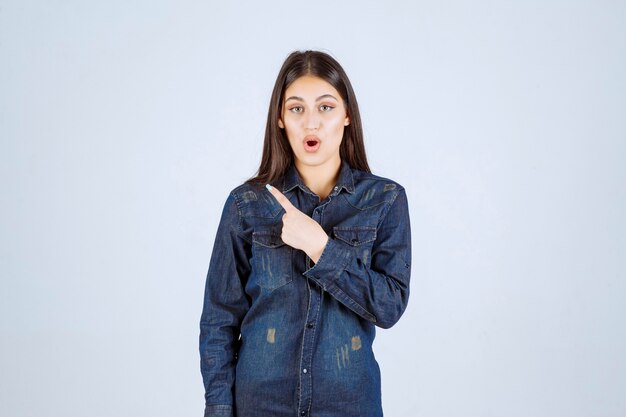 Jeune femme dans une chemise en jean pointant vers la gauche avec les émotions du visage