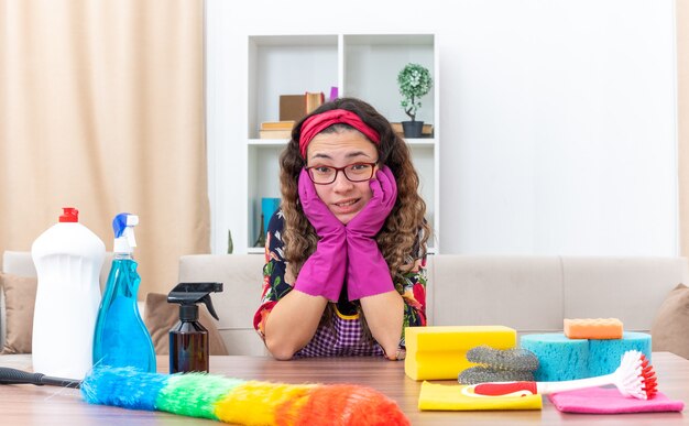 Jeune femme dans des gants en caoutchouc regardant la caméra confus assis à la table avec des produits de nettoyage et des outils