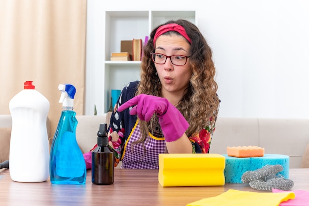 Photo gratuite jeune femme dans des gants en caoutchouc semblant étonnée et surprise assise à table avec des produits de nettoyage et des outils dans un salon lumineux