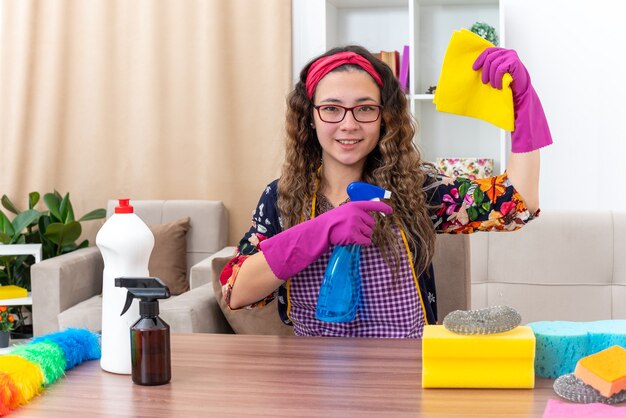 Jeune femme dans des gants en caoutchouc tenant un chiffon et un spray de nettoyage souriant heureuse et positive assise à la table avec des produits de nettoyage et des outils dans un salon clair