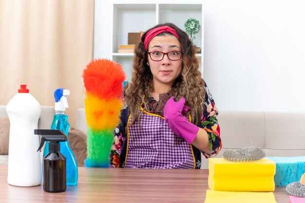 Jeune femme dans des gants en caoutchouc tenant un chiffon statique confus se pointant sur elle-même assise à la table avec des produits de nettoyage et des outils dans un salon clair