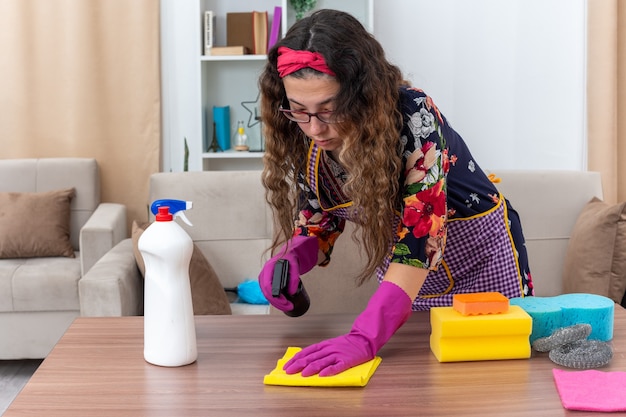 Jeune femme dans des gants en caoutchouc tenant un spray de nettoyage et une table de nettoyage de chiffon à l'aise dans un salon lumineux