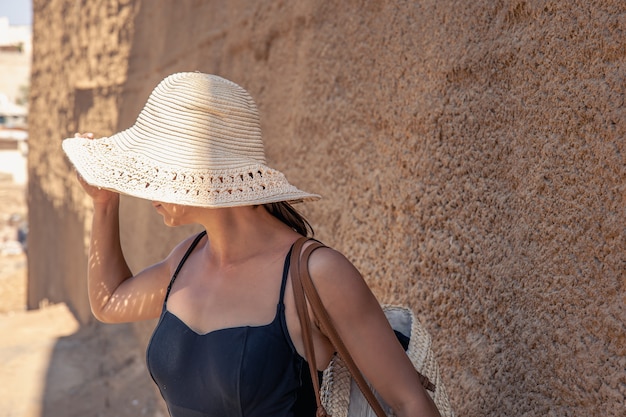 Une jeune femme dans un grand chapeau de paille se cache du soleil près d'un mur de sable par une chaude journée