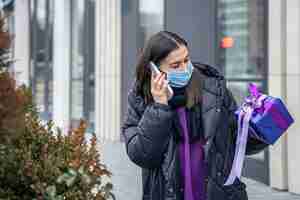 Photo gratuite jeune femme dans un masque de protection avec un cadeau dans ses mains parlant au téléphone