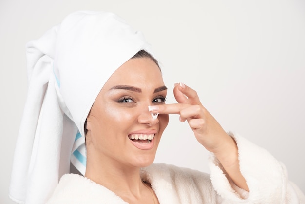 Jeune femme dans un peignoir blanc appliquant une crème sur son nez