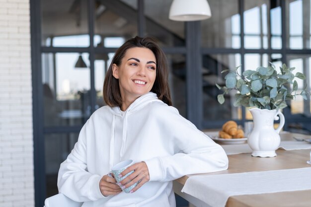 Jeune femme dans un sweat à capuche blanc chaud à la maison dans la cuisine, commence sa journée avec une tasse de café