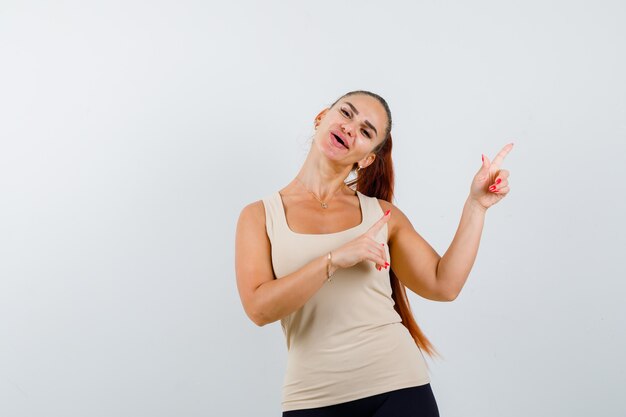 Jeune femme en débardeur beige pointant vers le coin supérieur droit et à la joyeuse vue de face.