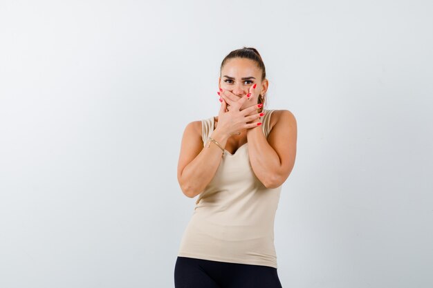 Jeune femme en débardeur beige tenant la main sur la bouche et à la perplexité, vue de face.