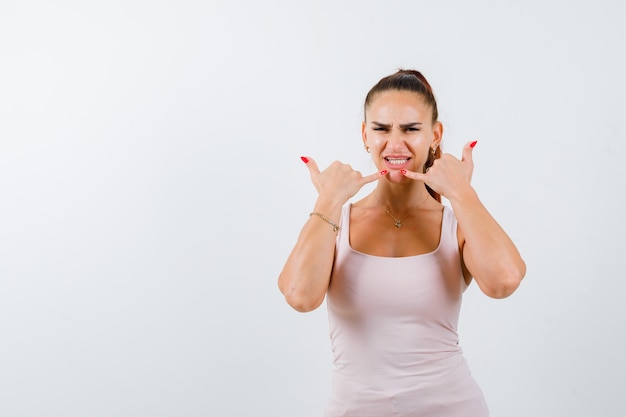 Jeune femme en débardeur blanc montrant le geste du téléphone et l'air confiant, vue de face.