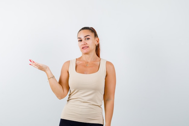 Jeune femme en débardeur faisant semblant de montrer quelque chose et à la vue indécise, de face.