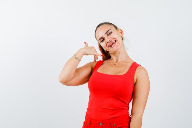 Jeune femme en débardeur rouge, haut de pantalon montrant le geste du téléphone et l'air heureux, vue de face.