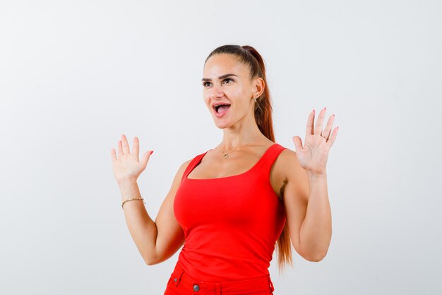 Jeune femme en débardeur rouge, pantalon montrant les paumes en geste de reddition et à la perplexité, vue de face.