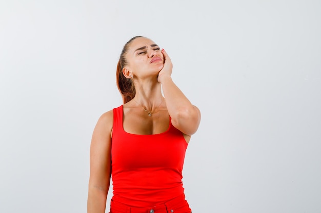 Jeune femme en débardeur rouge, pantalon souffrant de maux de dents et à la recherche de mal, vue de face.