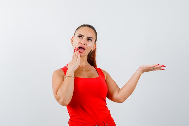 Jeune femme en débardeur rouge, pantalon tenant la main sur le menton tout en montrant quelque chose et à la perplexité, vue de face.